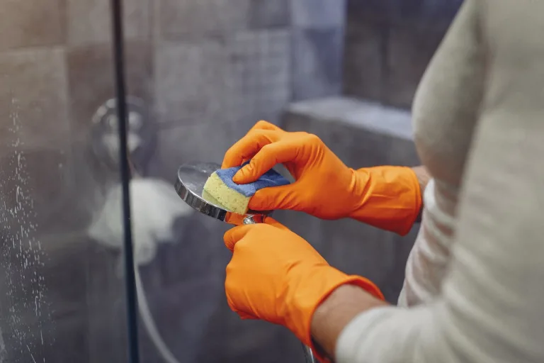 A person in orange gloves cleans a showerhead with a sponge in a tiled bathroom.
